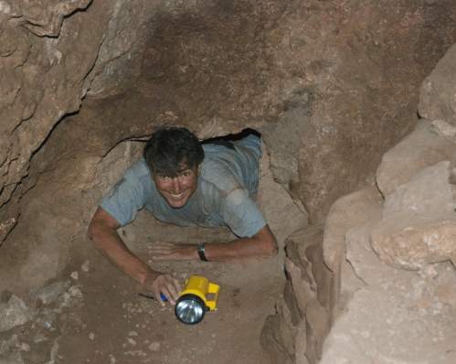 A person crawls through a narrow cave passage, holding a flashlight and smiling. Rocks surround the entrance.