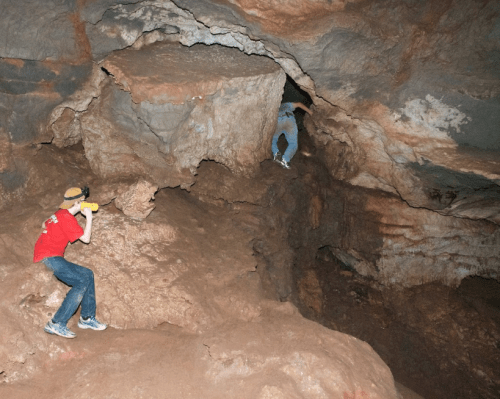 Two people exploring a cave, one climbing a rocky ledge while the other uses a flashlight to illuminate the area.