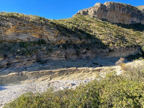 A rocky landscape with steep cliffs, sparse vegetation, and a dry riverbed under a clear blue sky.
