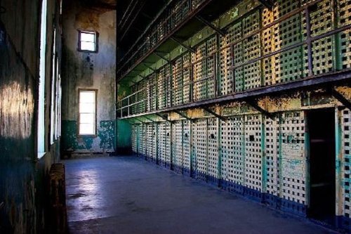 Abandoned prison corridor with peeling paint, empty cells, and sunlight streaming through tall windows.