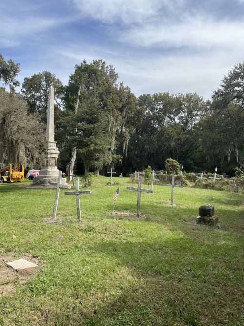 A cemetery scene featuring several wooden crosses, an obelisk, and lush greenery under a cloudy sky.
