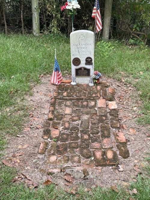 Grave of Alexander Lodge, marked by a stone headstone, flags, and flowers, surrounded by a brick pathway in a grassy area.