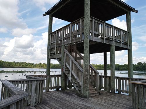 A wooden observation tower with stairs overlooking a serene lake surrounded by greenery and clouds.