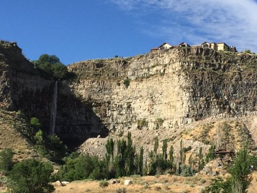 A rocky cliff with a waterfall cascading down, surrounded by greenery and houses perched on top against a blue sky.