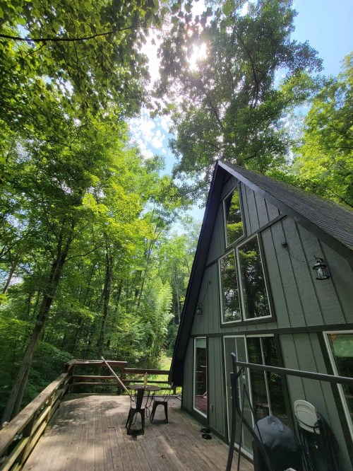 A-frame cabin nestled in lush green trees, with a wooden deck and bright sunlight filtering through the leaves.