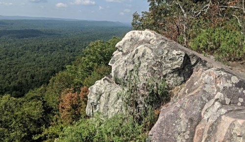 A rocky outcrop overlooks a lush green valley and distant mountains under a partly cloudy sky.