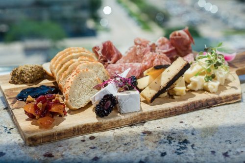 A wooden platter featuring assorted cheeses, cured meats, bread, and garnishes, set against a blurred city background.