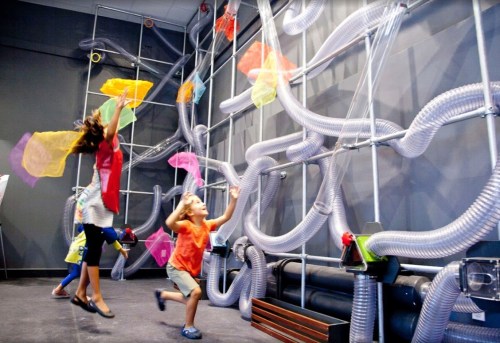Children play with colorful scarves in an interactive exhibit featuring tubes and flowing materials on a wall.