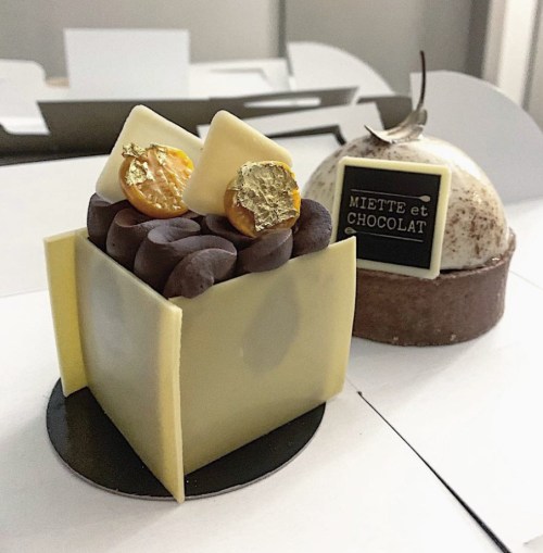 Two elegant desserts: a square chocolate cake topped with gold accents and a round cake with a decorative label.
