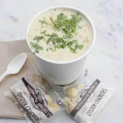 A cup of creamy soup topped with herbs, accompanied by two packets of oyster crackers on a napkin.