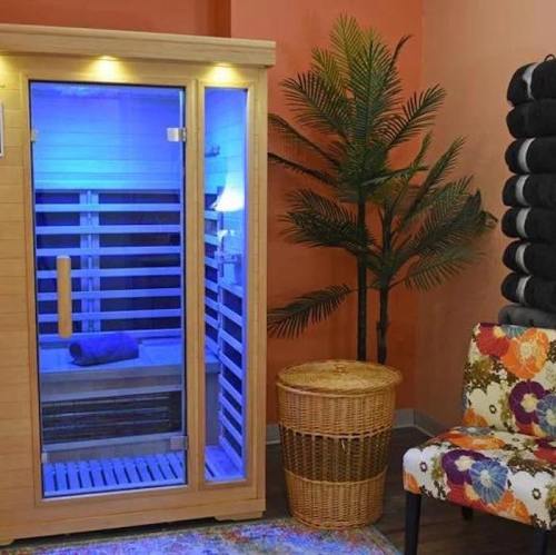 A wooden sauna with blue lighting, a towel inside, a potted plant nearby, and a colorful patterned chair.