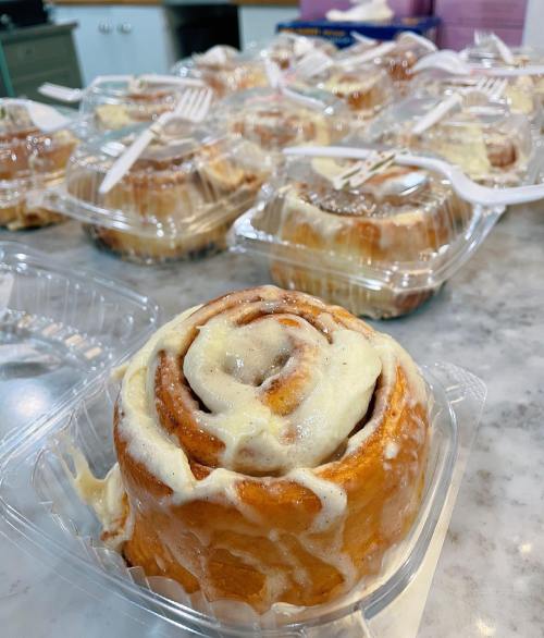 A close-up of a cinnamon roll with cream cheese frosting, surrounded by several more in clear plastic containers.