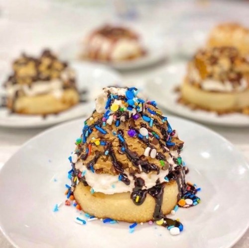 A close-up of a dessert topped with whipped cream, chocolate drizzle, and colorful sprinkles on a white plate.