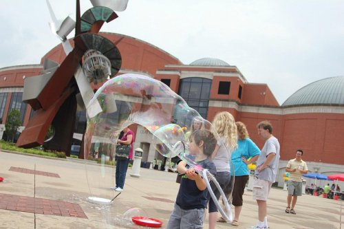 A child plays with a large bubble outside a building, with people and a sculpture in the background.
