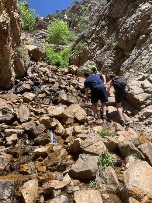 Two hikers navigate a rocky trail beside a small stream, surrounded by steep, rocky terrain and greenery.