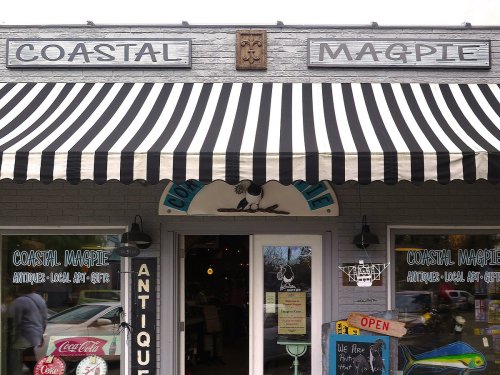 Storefront of "Coastal Magpie" with a striped awning, showcasing antiques and local art, featuring an "Open" sign.