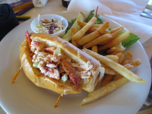 A lobster roll filled with lobster meat and celery, served with fries and a side of coleslaw on a white plate.