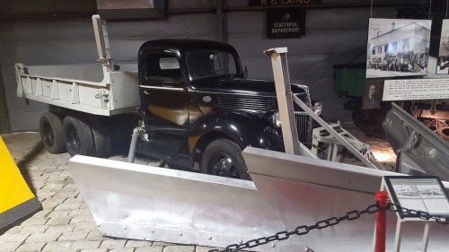 A vintage black truck with a flatbed and snow plow attachments displayed in a museum setting.