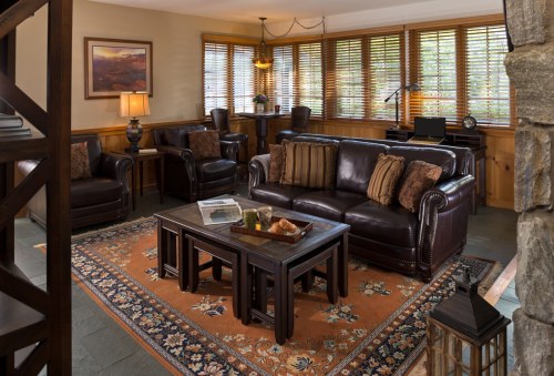 Cozy living room with leather sofas, wooden furniture, a patterned rug, and large windows with blinds.