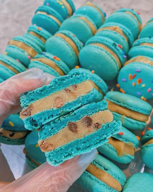 A hand holding a split turquoise macaron filled with cookie dough and chocolate chips, surrounded by more macarons.
