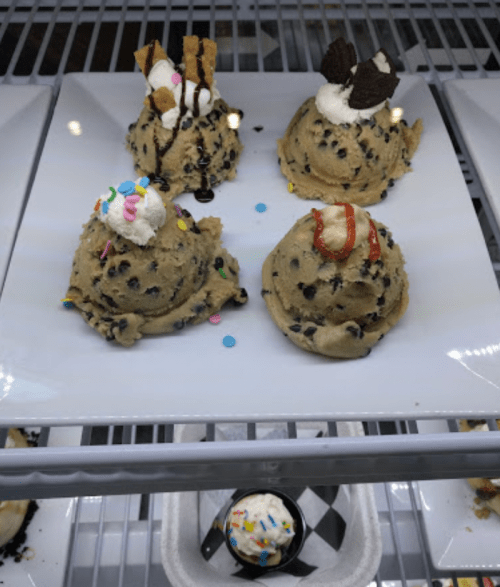 Four scoops of cookie dough with various toppings, including sprinkles, chocolate sauce, and whipped cream, on a white plate.