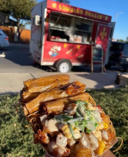 A hand holding a plate of crispy spring rolls topped with sauce and vegetables, with a food truck in the background.
