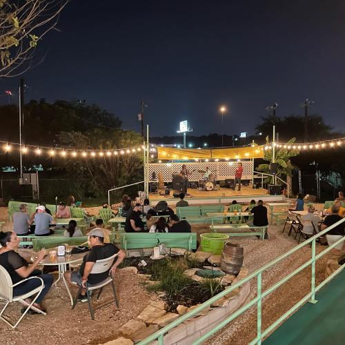 A lively outdoor venue at night with people seated, a stage, and string lights illuminating the area.