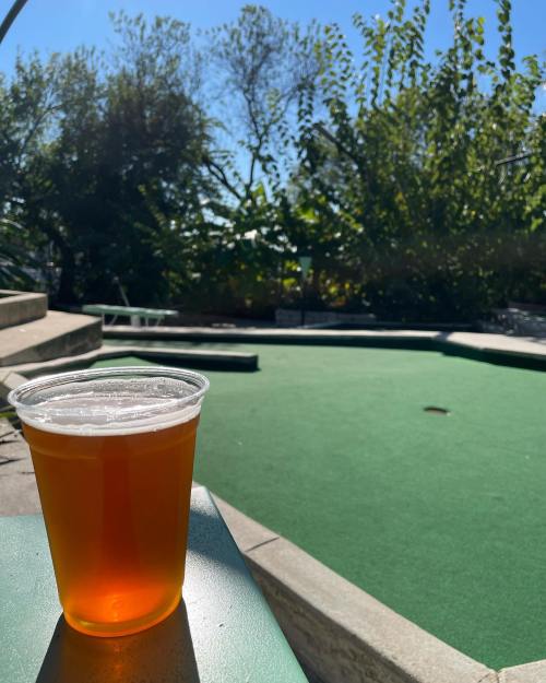 A clear plastic cup of beer sits on a table, with a mini-golf course and greenery in the background under a sunny sky.