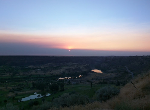 A serene sunset over a valley, with a river winding through lush greenery and soft clouds in the sky.