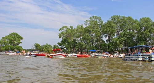 A busy lake scene with various boats and people enjoying a sunny day among green trees.