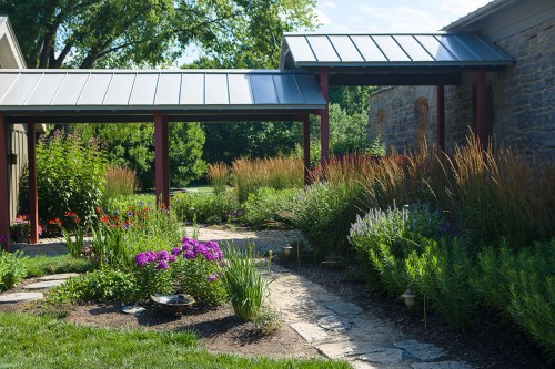 A vibrant garden with colorful flowers, stone pathways, and lush greenery, framed by modern architecture and sunlight.