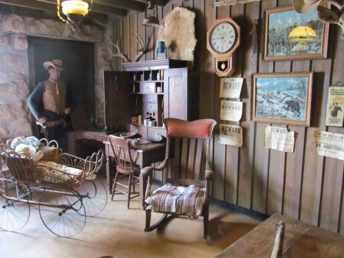 A rustic room with a wooden desk, rocking chair, vintage decor, and a mannequin dressed as a cowboy.