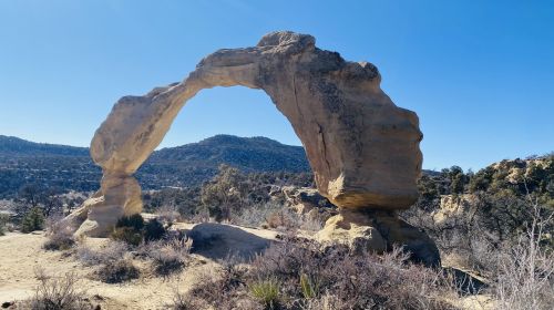 A natural stone arch stands against a clear blue sky, surrounded by rocky terrain and distant mountains.