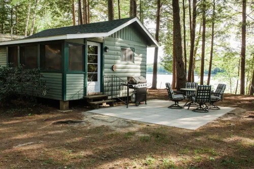 A cozy green cabin surrounded by trees, featuring a patio with a dining set and a grill near a lake.