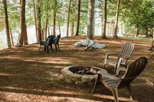 A serene outdoor scene with a fire pit surrounded by chairs, nestled among tall trees by a lake.