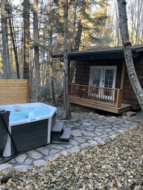 A cozy cabin surrounded by trees, featuring a hot tub on a stone path and a wooden deck.