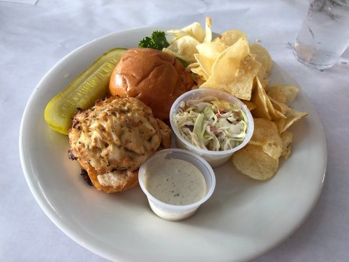 A plate with a crab cake sandwich, pickle, coleslaw, potato chips, and a side of dipping sauce.