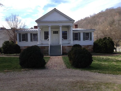A white, historic house with a porch, surrounded by greenery and a gravel path, set against a hilly backdrop.