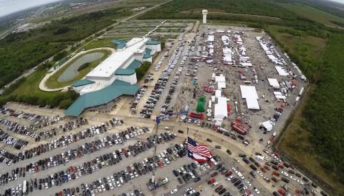 Aerial view of a large outdoor event with tents, cars parked, and green surroundings.