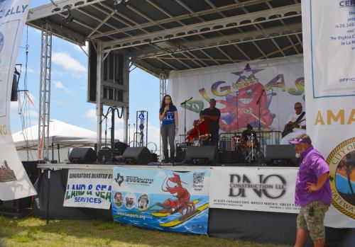 A singer performs on stage with a band at an outdoor event, surrounded by banners and a clear blue sky.