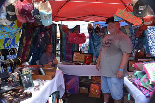 A vendor and a customer stand in a colorful market stall filled with jewelry, hats, and vibrant fabrics under a red canopy.