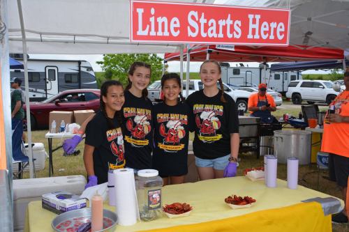 Four girls in matching shirts smile at a food booth with a sign that says "Line Starts Here" in a festival setting.