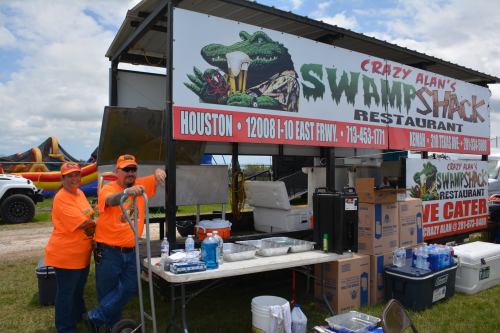 Two people in orange shirts stand by a food trailer with a sign for "Crazy Alan's Swamp Shack Restaurant."