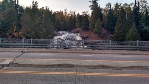A view of a waterfall surrounded by trees, seen from a bridge over a road.