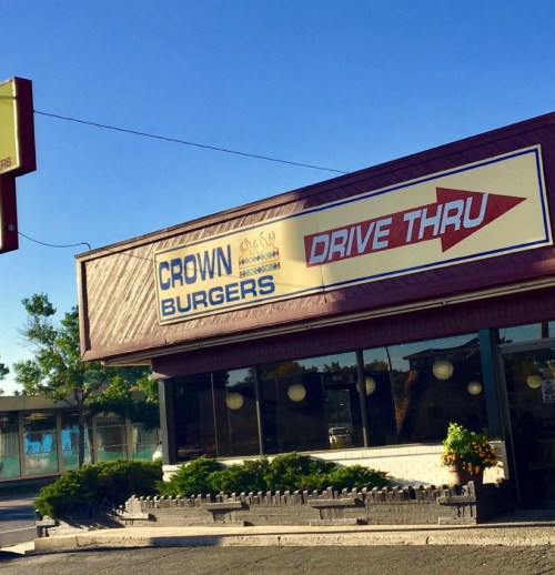 Sign for Crown Burgers with a drive-thru arrow, featuring a retro-style building and greenery in front.