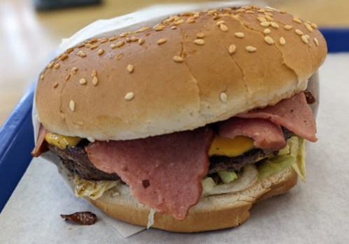 A close-up of a sesame seed bun burger with lettuce, cheese, and slices of deli meat on a tray.
