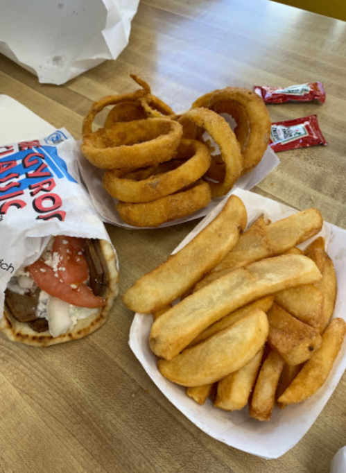A plate with a gyro sandwich, onion rings, and French fries, accompanied by two ketchup packets.