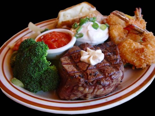 A plate featuring a grilled steak, shrimp, baked potato, broccoli, and dipping sauces.