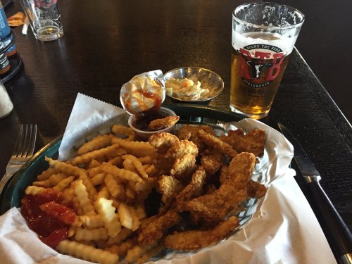 A plate of crispy chicken tenders with crinkle-cut fries, dipping sauces, coleslaw, and a pint of beer.