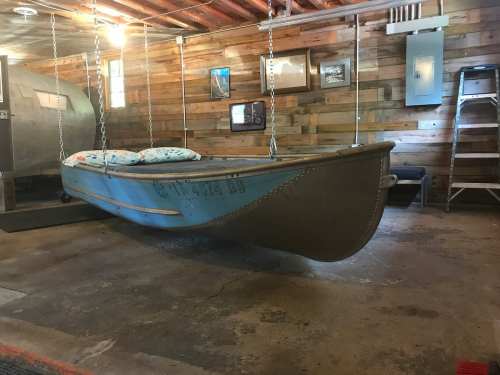 A hanging boat with cushions inside a rustic wooden room, featuring a concrete floor and framed pictures on the walls.